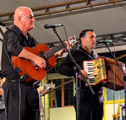 Dos hombres vestidos de negro presentan música en vivo sobre un escenario; el de la izquierda toca la guitarra acústica y el de la derecha toca el acordeón frente a un micrófono.