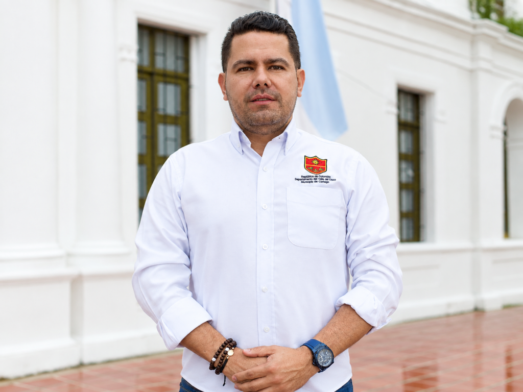 Persona de pie con camisa blanca de manga larga, sonriendo y posando frente a unas columnas y banderas en un entorno institucional.