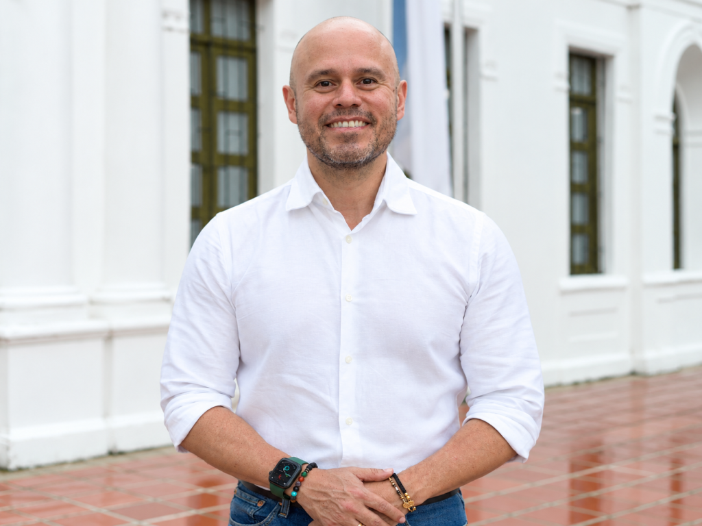 Persona de pie con camisa blanca de manga larga, sonriendo y posando frente a unas columnas y banderas en un entorno institucional.